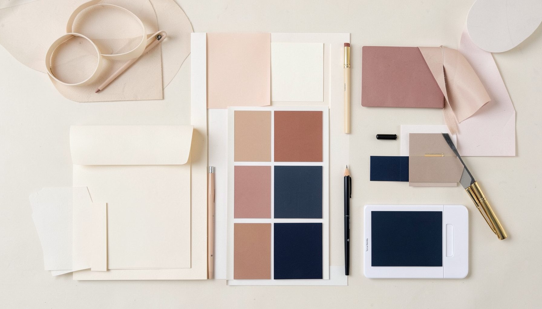 A tidy desk-level flat lay of design tools and color swatches in warm cream, dusty rose, and navy tones, arranged neatly to suggest a calm creative workspace